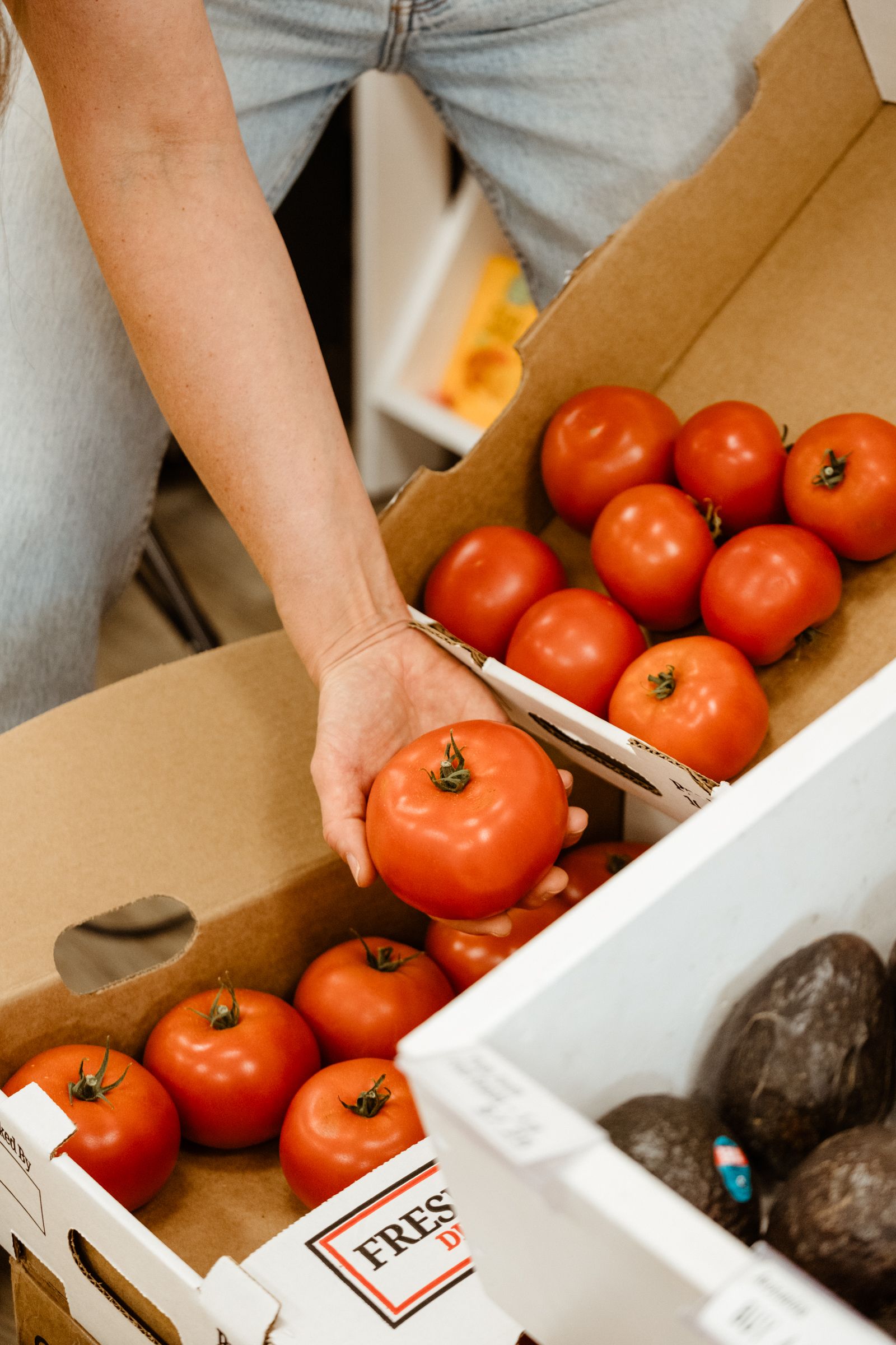 A hand selecting a tomato from crates of produce, with avocados nearby