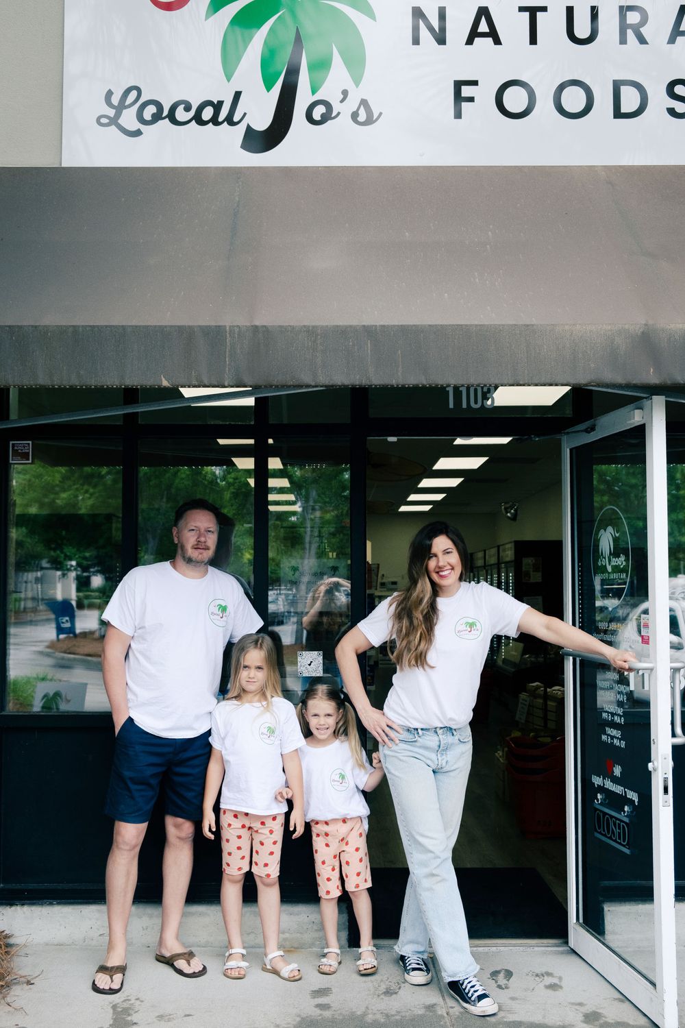 Jo and Chad standing with their two daughters outside the Local Jo's Natural Foods storefront, all wearing matching store T-shirts